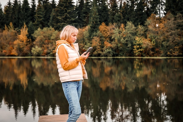 Happy young pretty woman walking outdoors in autumn park using mobile phone chatting scenic view of the river