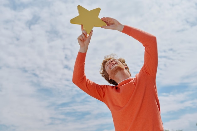 Happy young redhead man holding yellow star and smiling with sky in the background