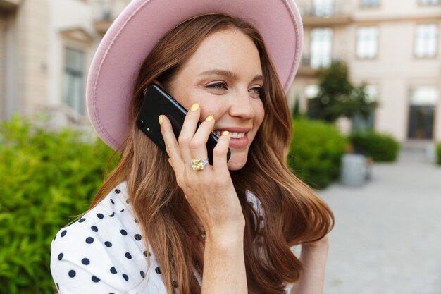 happy young redhead woman sit on a bench outdoors at the street talking by mobile phone.