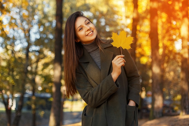 Happy young smiling woman in a fashionable coat with a yellow autumn leaf walks in the park at sunset