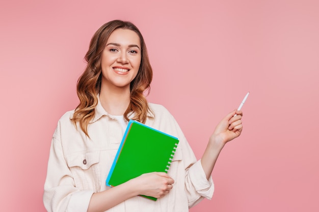 Happy young student girl smiles and points a finger at empty . Portrait of a young caucasian woman with a pen and notobook notepad isolated over pink wall business freelancer