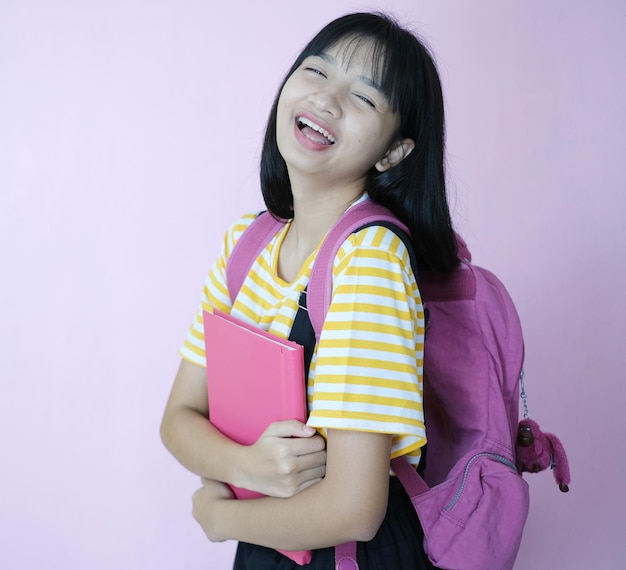 Happy young student holding pink book with backpack on pink background.