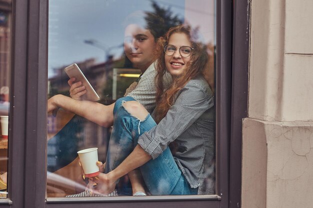 Happy young students drinking coffee and using a digital tablet sitting on a window sill at a college campus during a break.