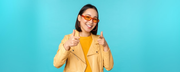 Happy young stylish chinese girl in sunglasses points fingers at camera with pleased smile choosing