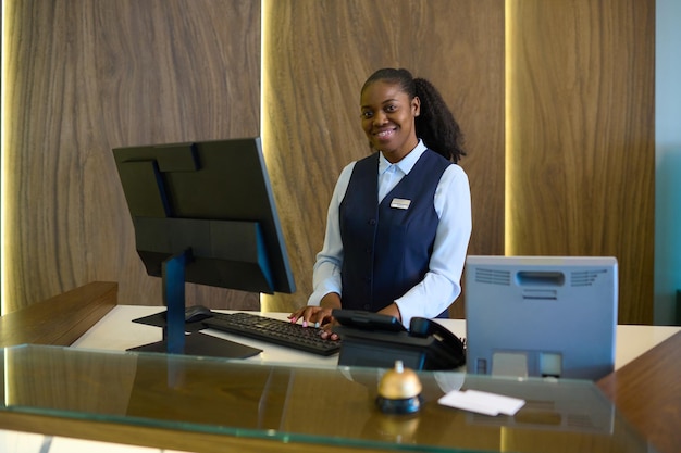 Happy young successful female receptionist in uniform standing by counter