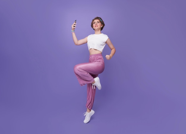 Happy young teen woman celebrating with mobile phone while jumping up on studio background full body