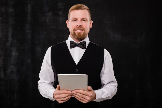 Happy young waiter in bowtie and black waistcoat holding digital tablet while standing in front of camera