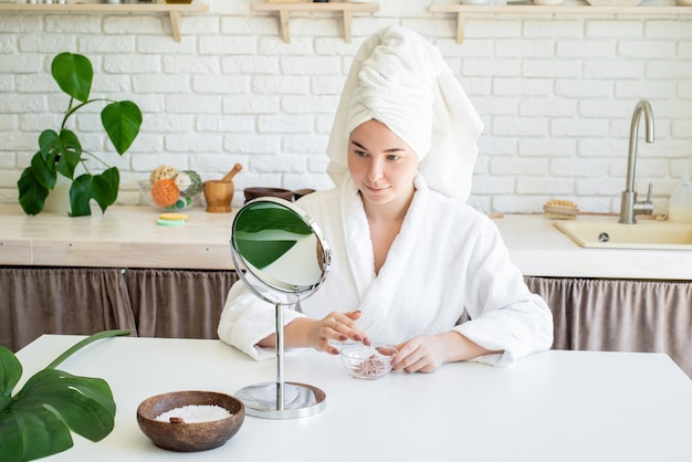 Happy young woman applying face scrub on her face in her home kitchen looking at the mirror