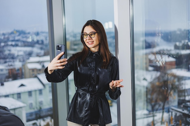 Happy young woman in a black shirt smiling and talking on the phone in a modern office with large windows Remote work