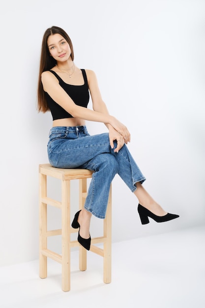 Happy young woman in black tank top and wide jeans in profile on chair