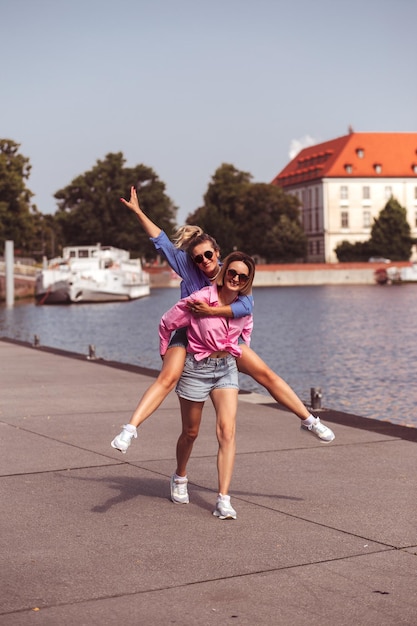 Happy young woman blond hung on back of her friend against the open background of city Girls are smiling broadly at camera