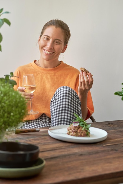 Happy young woman in casual clothes smiling and looking at camera while eating tasty steak tartare with crouton and drinking wine at home