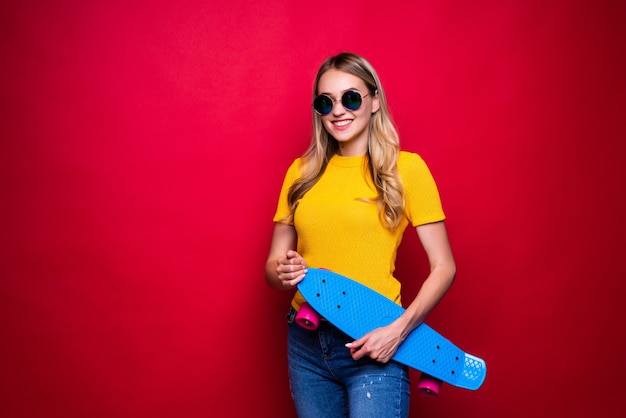 Happy young woman in casual outfit and hat carrying skateboard on shoulder  against red wall