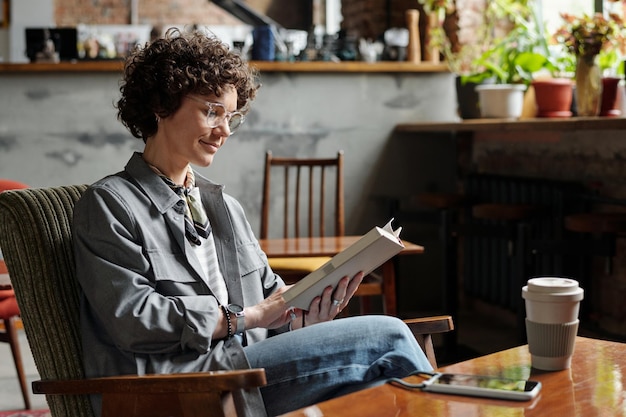 Happy young woman in casualwear and eyeglasses relaxing in armchair with book