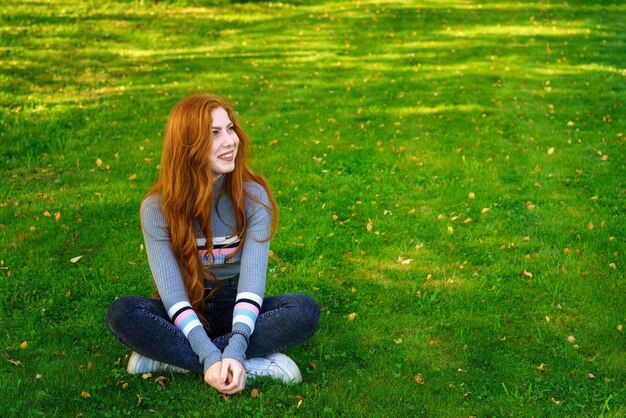 Photo happy young woman of caucasian ethnicity with long red hair sits in casual clothes in the park on green grass smiling and looking to the side on a sunny day