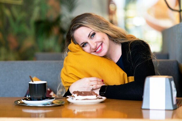 Photo happy, young woman in a coffee shop hugging a pillow. the female smiles and drinks coffee, cheerfulness in the morning