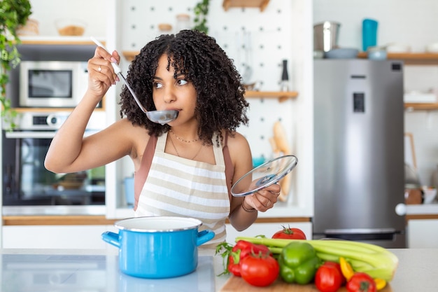 Happy Young Woman Cooking Tasting Dinner In A Pot Standing In Modern Kitchen At Home Housewife Preparing Healthy Food Smiling Household And Nutrition Dieting Recipes Concept