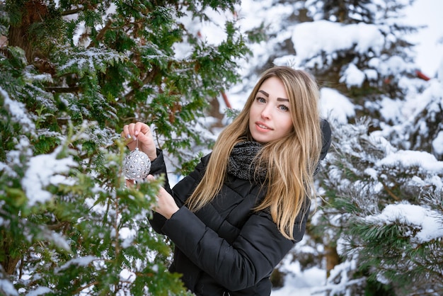 Happy young woman decorating pine branches with modern bauble decorating christmas tree in snow outd...