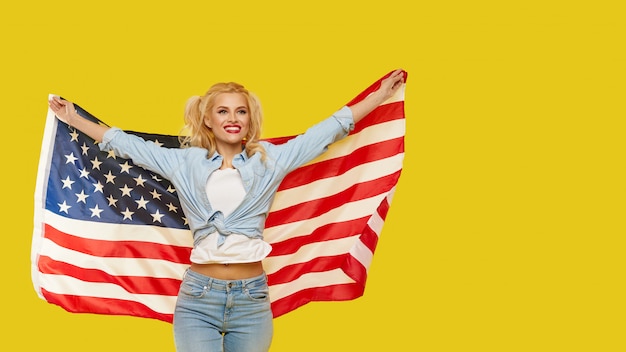 Happy young woman in denim clothes holding USA flag isolated on yellow background