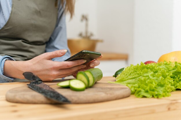 Happy young woman drinking orange juice, browsing social networks on her smartphone while cooking. Close up.