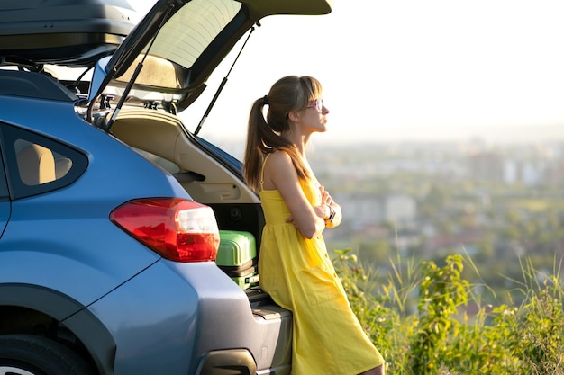 Happy young woman driver in yellow dress enjoying warm summer evening standing beside her car