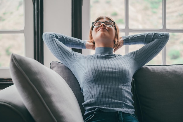 Happy young woman in eyeglasses relaxing on sofa with hands behind head in the living room of her house Carefree and satisfied lady with relaxing on couch at modern apartmentxA