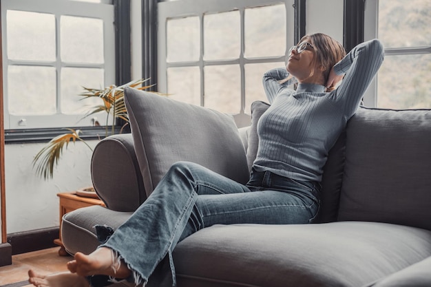 Photo happy young woman in eyeglasses relaxing on sofa with hands behind head in the living room of her house carefree and satisfied lady with relaxing on couch at modern apartmentxa