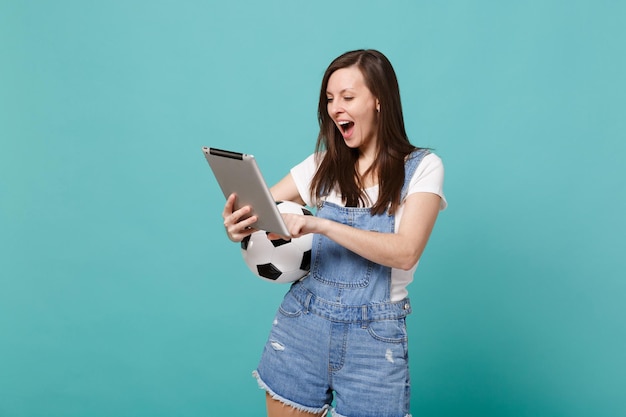 Happy young woman football fan holding soccer ball, using tablet pc computer isolated on blue turquoise wall background. People emotions, sport family leisure lifestyle concept. Mock up copy space.