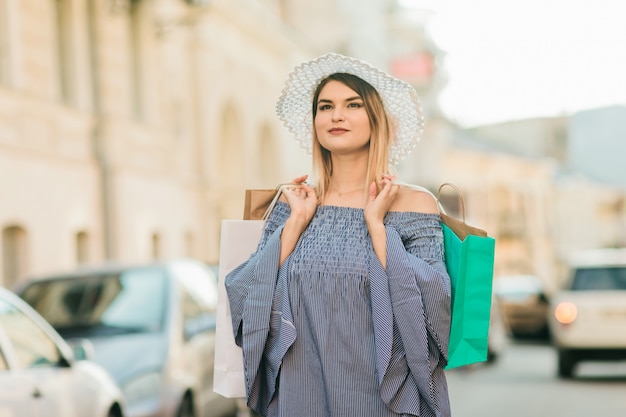 Happy Young woman in a hat and dress holds shopping bags in her hands and smiles in a summer city