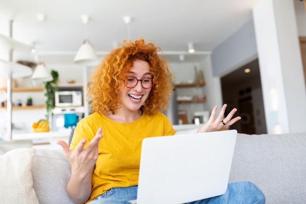 Happy young woman having fun doing video call using laptop in her home waving hand video conference calling on laptop computer sit on sofa distance learn zoom online virtual meeting at home