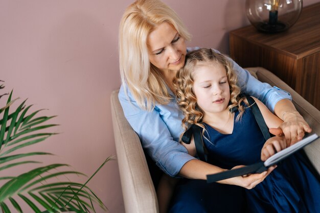 Photo happy young woman and her little curly daughter reading book together while sitting on armchair