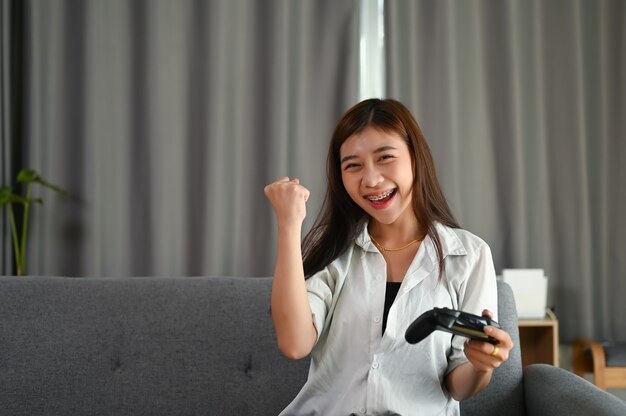 Happy young woman is playing and winning online game while sitting on sofa at home.