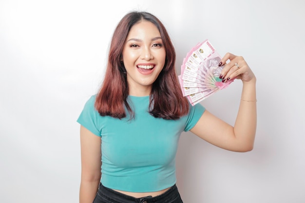 A happy young woman is wearing blue tshirt and holding cash money in Indonesian rupiah isolated by white background