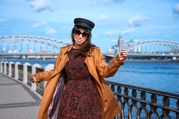 Happy young woman in a light brown coat and black cap posing on the embankment against the backdrop of the bridge and the blue sky