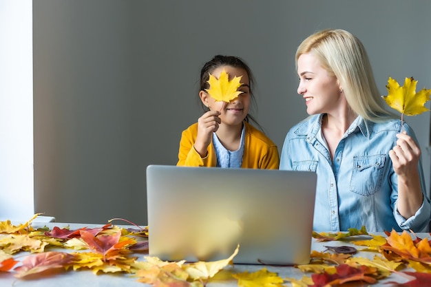 Happy young woman and little girl working online, watching webinar, podcast on laptop, having remote conversation at home around autumn leaves