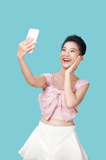 Happy young woman making silly faces while taking selfie on smartphone app with filters standing against blue background