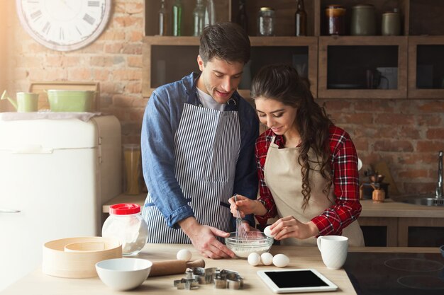 Happy young woman and man baking pie in loft kitchen. Young family cooking at home, using digital tablet. Mockup for recipe