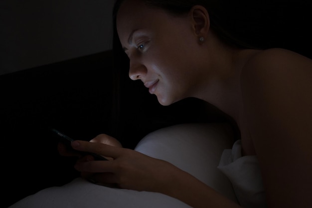 Happy young woman messaging on mobile phone while lying in soft bed late at night at home