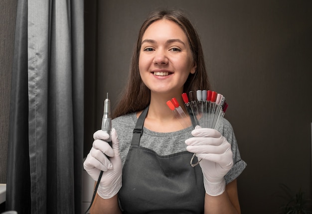 Happy young woman profession manicurist with nail machine and nail color palette on gray background