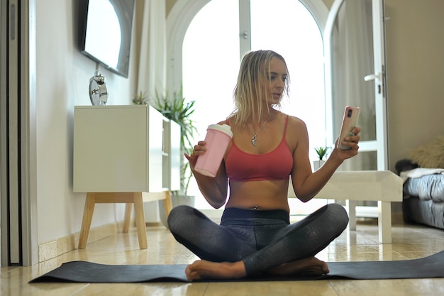 Happy young woman resting on yoga mat drinking water and checking phone