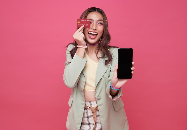 Happy young woman showing credit card and mobile phone standing over pink background