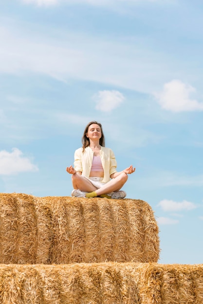 Happy young woman sits in lotus position on huge haystack and meditates Blue sky background Countryside holiday