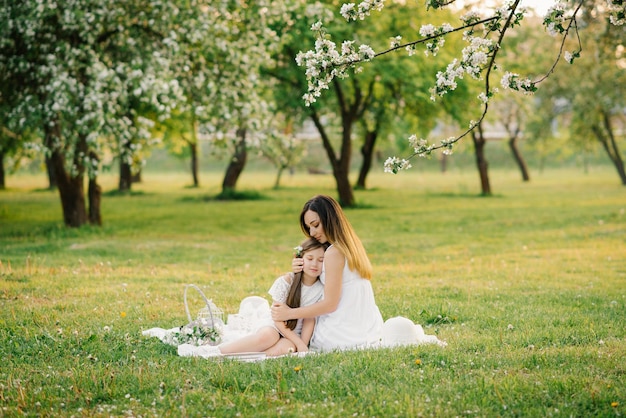 A happy young woman sits on a picnic blanket and hugs her daughter mother and daughter's summer picnic in the apple orchard