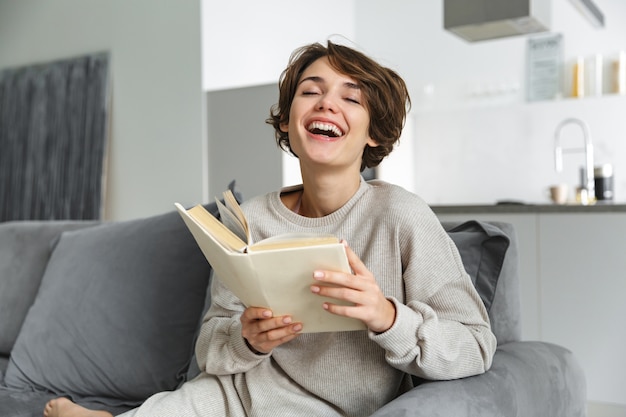 Happy young woman sitting on a couch at home, reading a book