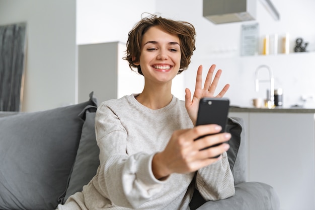 Happy young woman sitting on a couch at home, using mobile phone, making a video call