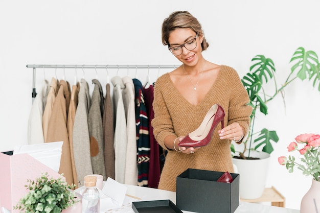 Happy young woman in smart casual knitted pullover standing by desk while holding fashionable shoe on high heel