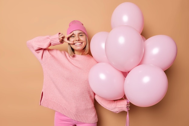 Happy young woman smiles broadly makes peace gesture over eye enjoyss life wears casual jumper and pink hat holds bunch of inflated balloons isolated over brown background Festivity concept