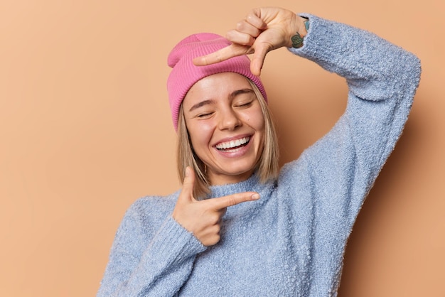 Happy young woman smiles broadly tries to find perfect angle makes frame gesture over face with fingers makes memorable photo wears pink hat casual blue jumper poses against brown background