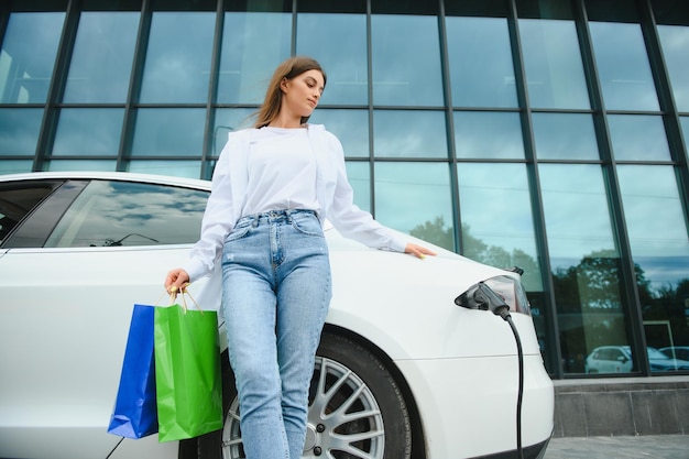 Happy young woman standing on city parking near electric car charging automobile battery from small city station holding shopping bags