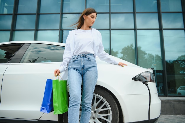 Happy young woman standing on city parking near electric car charging automobile battery from small city station holding shopping bags